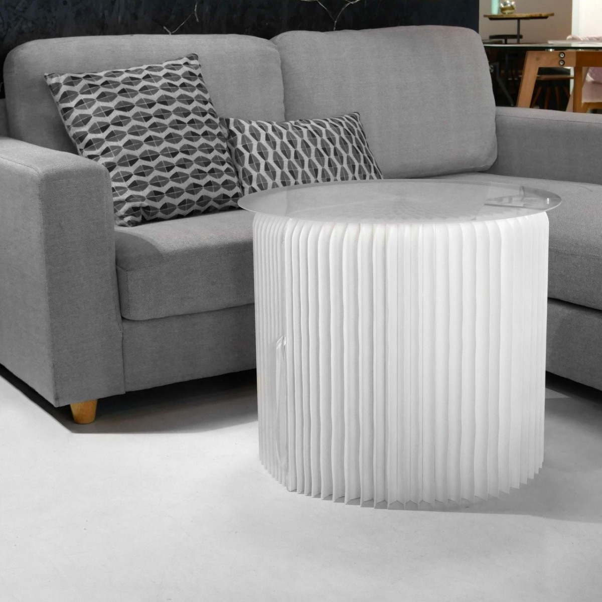 Table basse blanche en carton alvéolé posée à côté d'un canapé pour compléter le salon, avec un plateau e plexiglass pour poser des objets.